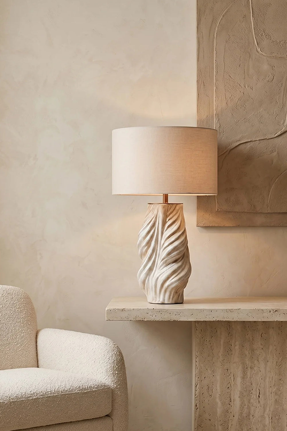 Adara Sculptural Ceramic Table Lamp | Ceramic, Fabric | Displayed on a modern floating travertine ledge next to a cream armchair against a minimalist textured plaster wall nook | vexa lamps