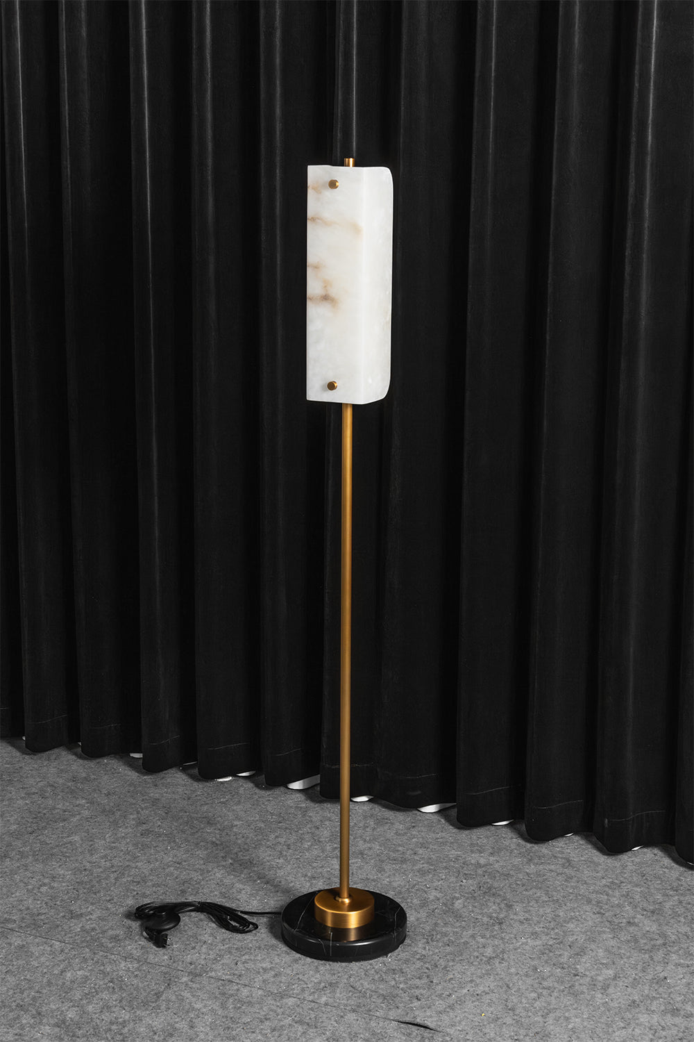Aurelius Alabaster Brass Floor Lamp | Alabaster, Brass | Staged studio shot of the lit alabaster standing light radiating a warm yellow halo on a grey carpet against black vertical drapes | vexa lamps