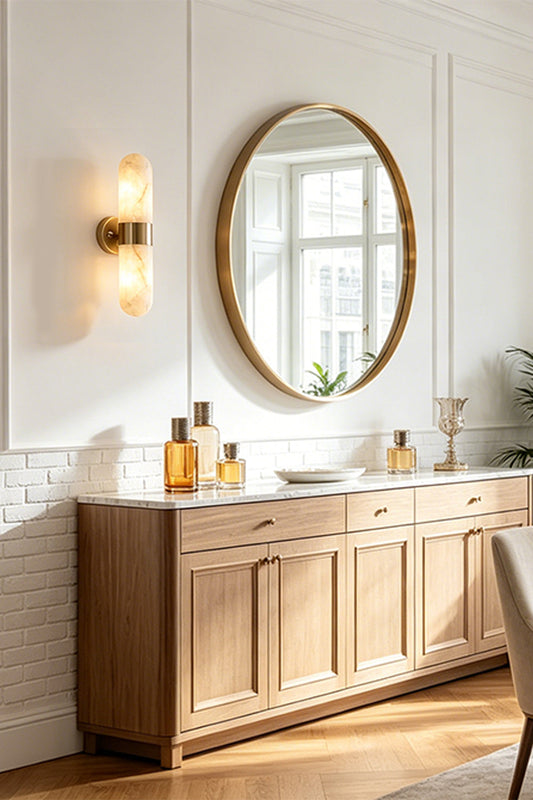 Calla Alabaster Capsule Wall Sconce | Alabaster, Brass | Mounted elegantly on a white wall next to a large round gold mirror and a wooden sideboard cabinet in a luxury hallway | vexa lamps