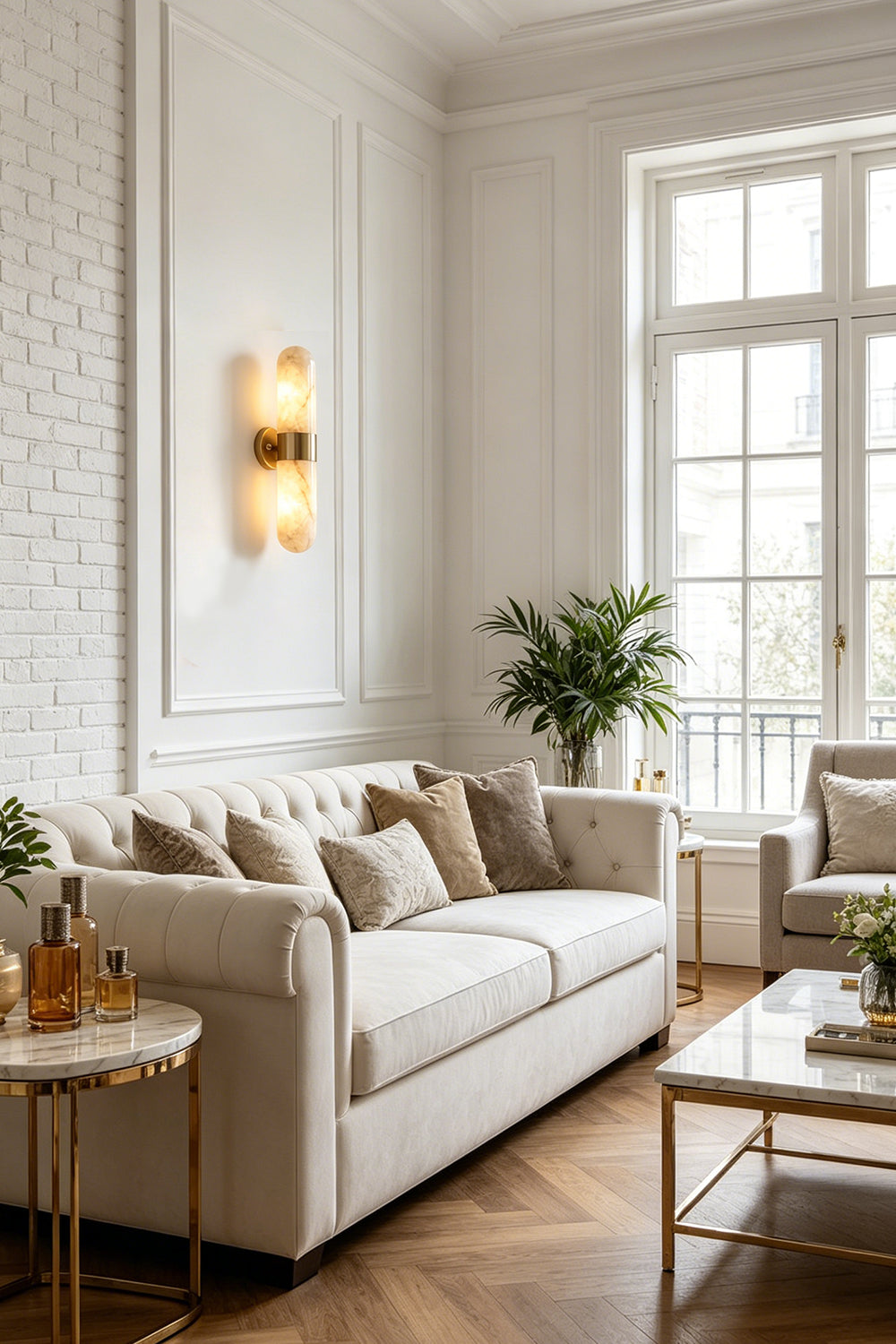 Calla Alabaster Capsule Wall Sconce | Alabaster, Brass | Providing a warm ambient glow on a white paneled wall in a sophisticated living room next to a cream fabric sofa | vexa lamps