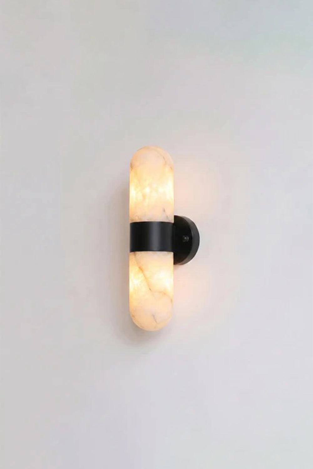 Calla Alabaster Capsule Wall Sconce | Alabaster, Metal | Studio front view of the fully lit matte black hardware variant casting a warm yellow halo against a plain background | vexa lamps