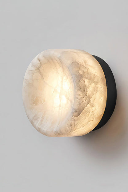 Isolde Concave Alabaster Disc Wall Sconce | Natural Alabaster and Black Metal | Studio close-up shot of the lit sconce featuring a matte black finish base on a light wall | vexa lamps