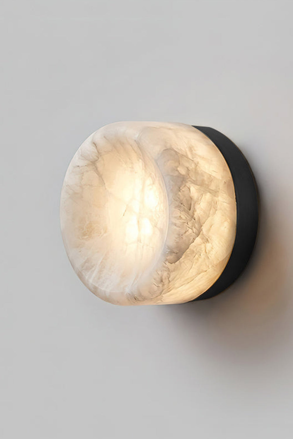 Isolde Concave Alabaster Disc Wall Sconce | Natural Alabaster and Black Metal | Studio close-up shot of the lit sconce featuring a matte black finish base on a light wall | vexa lamps