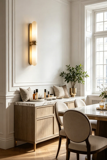 Linea Alabaster Capsule Wall Sconce | Alabaster, Brass | Radiating warm ambient lighting on a white wall near a dining area with a classic sideboard and chairs | vexa lamps