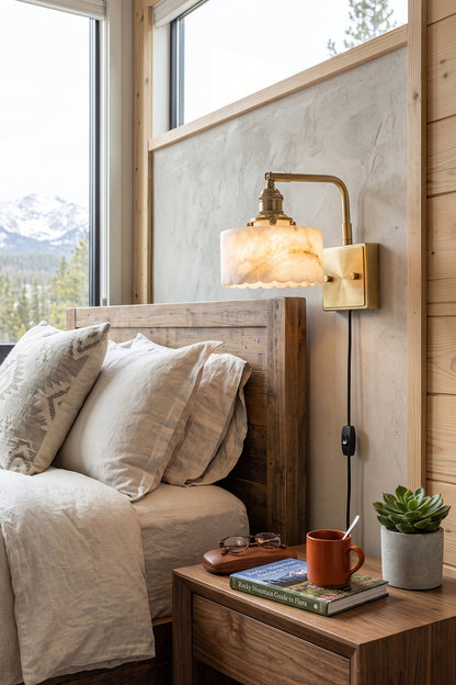 Liora Scalloped Alabaster Wall Sconce | Natural Alabaster and Brushed Brass | Mounted on a wooden bedroom wall beside a bed and a rustic nightstand with a snowy mountain window view | vexa lamps