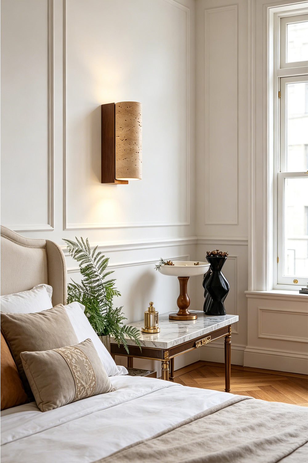 Outcrop Beige Travertine Wall Sconce | Travertine, Metal | Mounted as an elegant bedside accent light casting a warm upward and downward glow on a white wall next to a bed with a cream headboard | vexa lamps