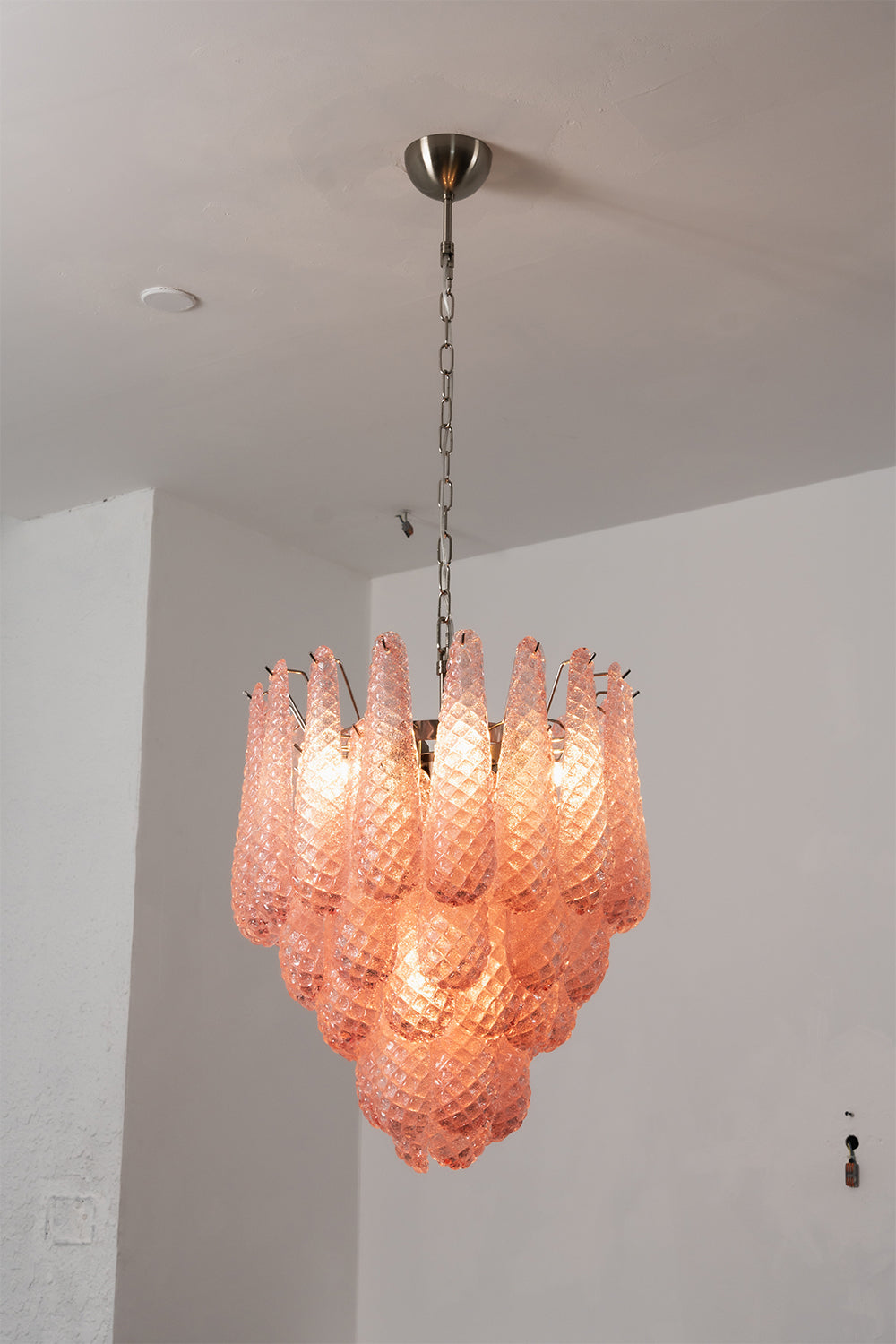 Amara Blush Cascading Glass Chandelier | Glass, Metal | Close-up frontal view of the glowing blush pink cascading chandelier against a plain grey wall | vexa lamps