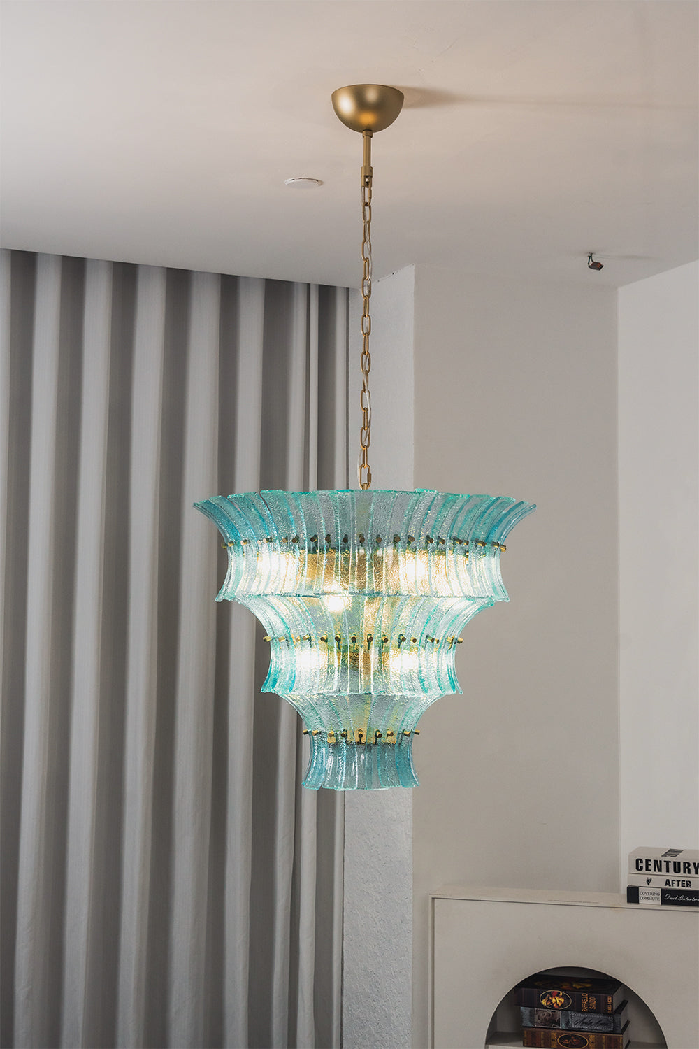 Thalassa Aquamarine Textured Glass Chandelier | Glass, Metal | Alternative studio shot of the illuminated tiered architectural glass light fixture hanging elegantly next to a grey drapery panel | vexa lamps