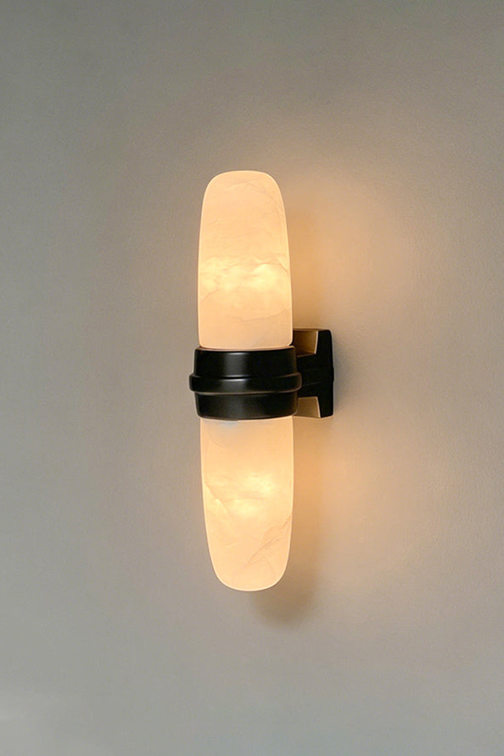 Theron Linear Alabaster Wall Lamp | Alabaster, Metal | Vertical shot of the illuminated alabaster sconce with a dark black center band on a plain grey backdrop | vexa lamps