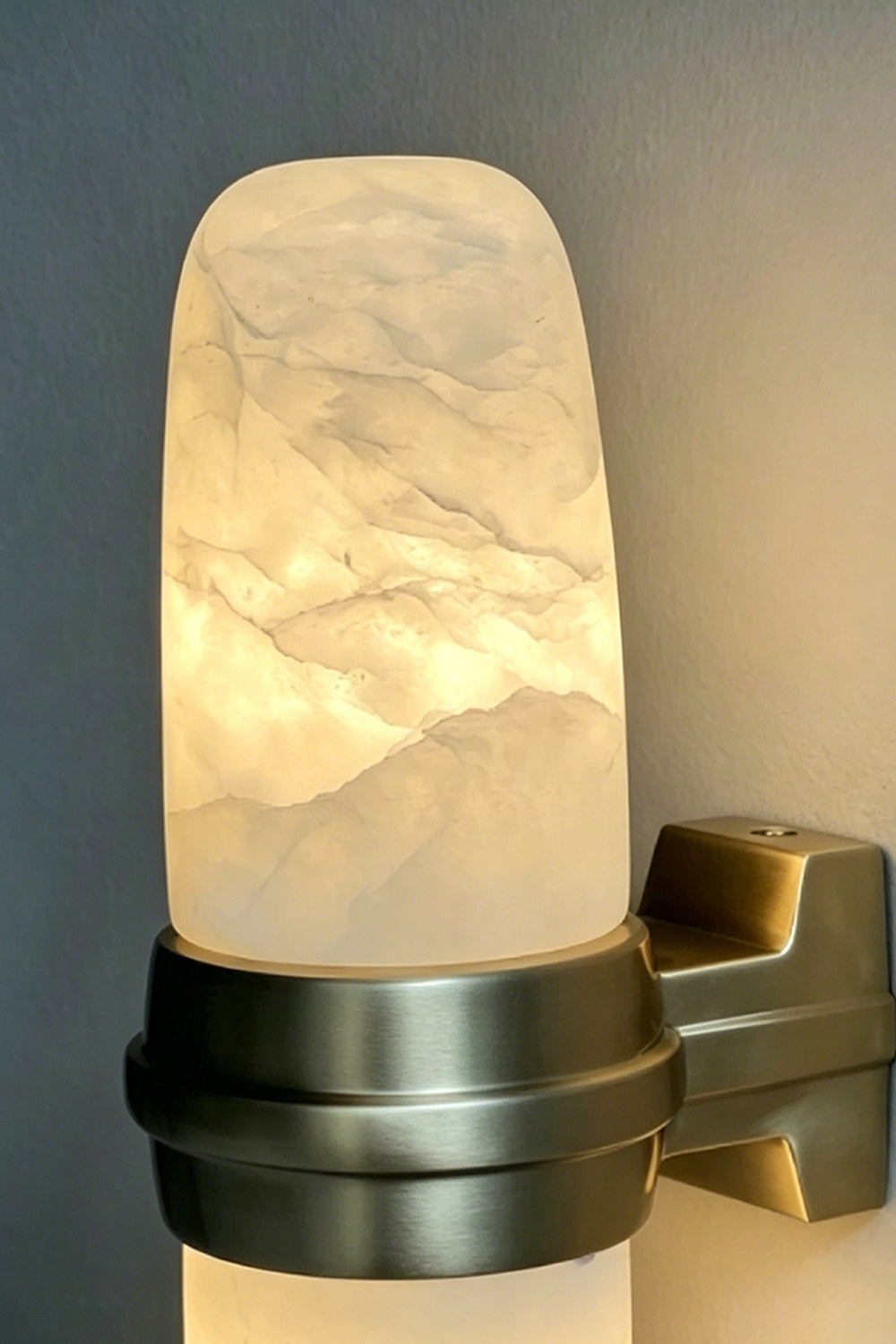 Theron Linear Alabaster Wall Lamp | Alabaster, Metal | Moody close-up focus on the dark bronze mounting band holding the glowing translucent stone capsule under low light | Detail shot | vexa lamps